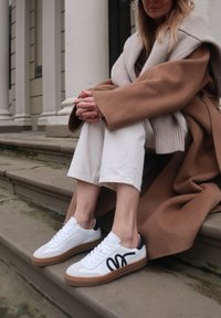 Person in beige coat and white pants sitting on stone steps wearing white sneakers with black design and gum soles.