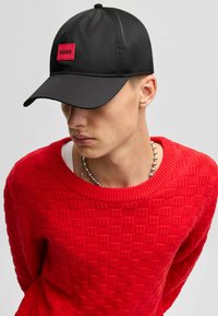 Sort kasket med flad skygge, der har et rødt rektangulært logo mærke med teksten "HUGO." Glat overfladetextur, sammen med en model i en rød mønstret sweater.