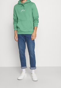Hombre que lleva puesta una sudadera verde, jeans azules remangados, calcetines blancos y zapatillas blancas, de pie contra un fondo sencillo.