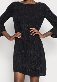 Vestido negro con un patrón geométrico, con mangas tres cuartos y cuello redondo. Tejido texturizado con toques brillantes.