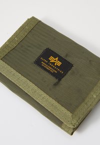 Portefeuille vert olive en tissu texturé, ornée d'un label noir avec un logo jaune et des bords cousus. Forme compacte et rectangulaire.