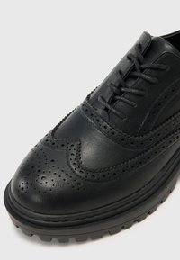 Chaussure brogue en cuir noir avec des détails perforés, un bout arrondi et une semelle en caoutchouc épaisse et texturée. Les lacets passent à travers cinq œillets.