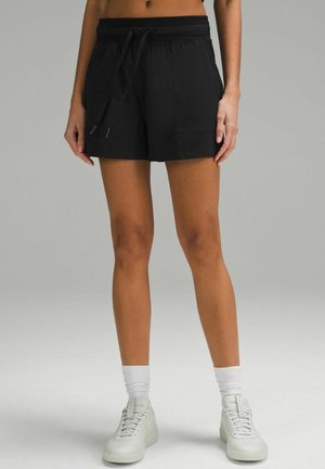 Persona con pantalones cortos negros con cordón, calcetines blancos hasta la pantorrilla y zapatillas blancas de pie contra un fondo gris liso.