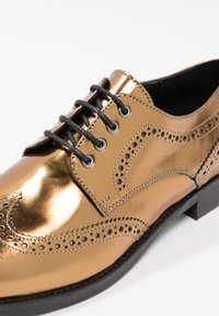 Chaussure brogue en cuir bronze brillant avec lacets noirs, perforations décoratives et semelle noire, présentée en angle pour montrer les détails avant et latéraux.