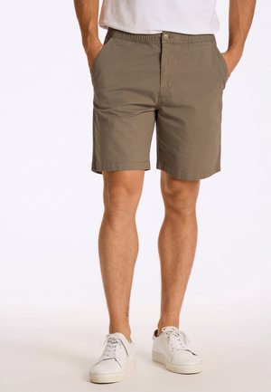 Mand iført olivengrønne shorts, hvide sneakers og en hvid skjorte, stående med hænderne i lommerne på en hvid baggrund.