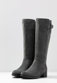 Stivali alti in suede grigio con punta tonda, zip laterale e una piccola fibbia decorativa sulla parte superiore. Tacco piatto e texture liscia.