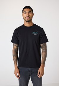 Svart bomull T-skjorte med rund hals og korte ermer, med turkis og hvit tekstlogo på venstre brystområde.