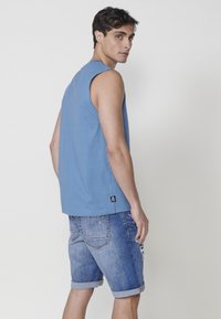 Modra brezrokavna majica iz bombaža, v kombinaciji z svetlo modrimi uničenimi denim kratkimi hlačami. Vidna obroba in detajl logotipa na hrbtni strani kratkih hlač.