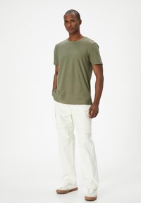 Camiseta verde oliva de tela suave, combinada con pantalones cargo blancos de corte holgado y zapatillas de colores claros. Diseño sencillo y casual.