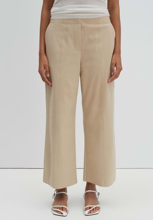 Personne portant un pantalon ample beige et des sandales blanches à brides, debout devant un fond clair uni.