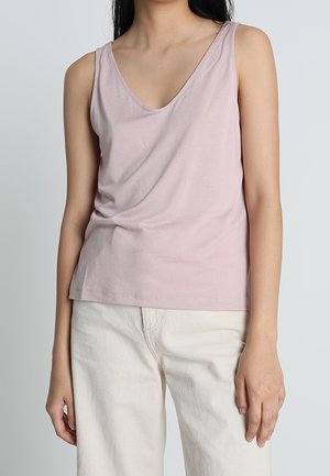 Kobieta w jasnoróżowym, bezrękawnym topie z okrągłym dekoltem i kremowych spodniach na jednolitym szarym tle.