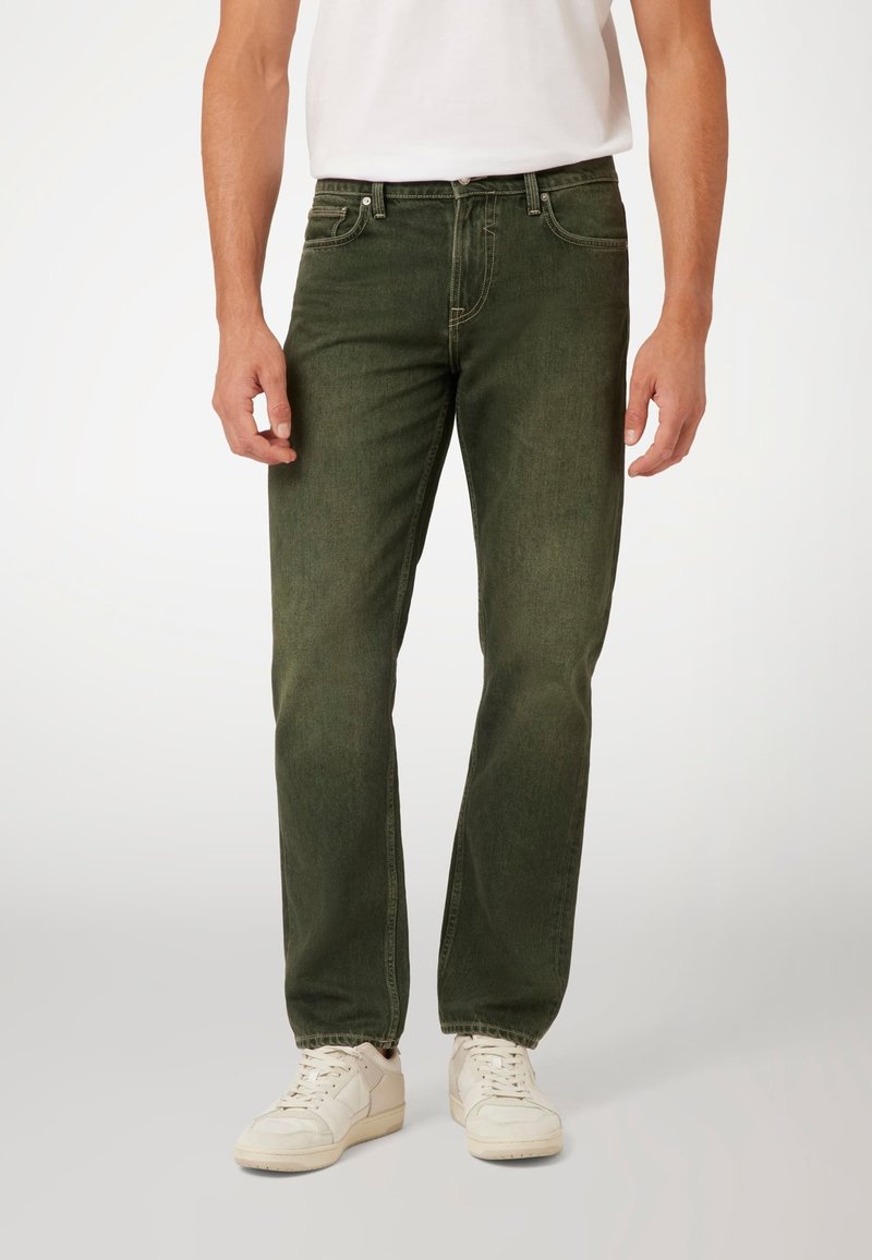 Alyvuogių spalvos džinsai su tiesiu kirpimu, penkiomis kišenėmis ir matomomis siūlėmis. Dėvimi su baltais sportiniais bateliais ir paprastu baltu marškinėliu.