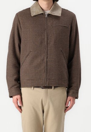 Homme portant une veste zippée marron à motif pied-de-poule avec un col en velours côtelé beige et un pantalon beige, debout sur fond blanc.