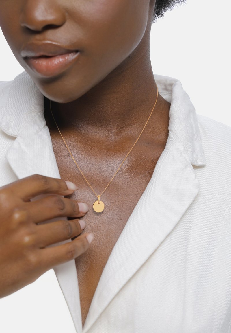 Gouden ketting met een ronde hanger, gedragen op gebruinde huid, met een gestructureerde witte stof met kraag op de achtergrond.