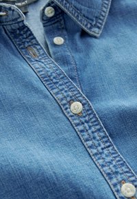 Jeanshemd in Hellblau mit klassischem Knopfverschluss, weißen Knöpfen und sichtbaren Nähten entlang des Kragens und der Knopfleiste.
