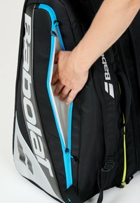 Mano che apre la tasca con zip di una borsa sportiva Babolat nera e grigia con rifiniture blu.