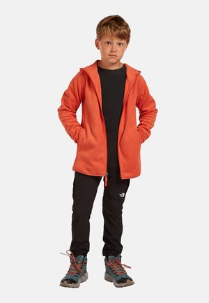 Niño de pie con las manos en los bolsillos, vistiendo una sudadera con capucha naranja con cremallera, camiseta negra, pantalones negros y botas de senderismo grises con cordones naranjas.