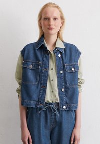 Denim vest in donkerblauw met twee voorzakken, zilveren drukknoopjes en een cropped design, gelaagd over een lichtgroen shirt.