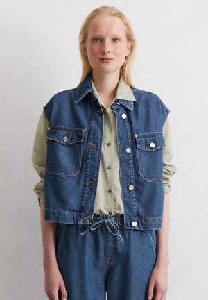 Denim vest in donkerblauw met twee voorzakken, zilveren drukknoopjes en een cropped design, gelaagd over een lichtgroen shirt.