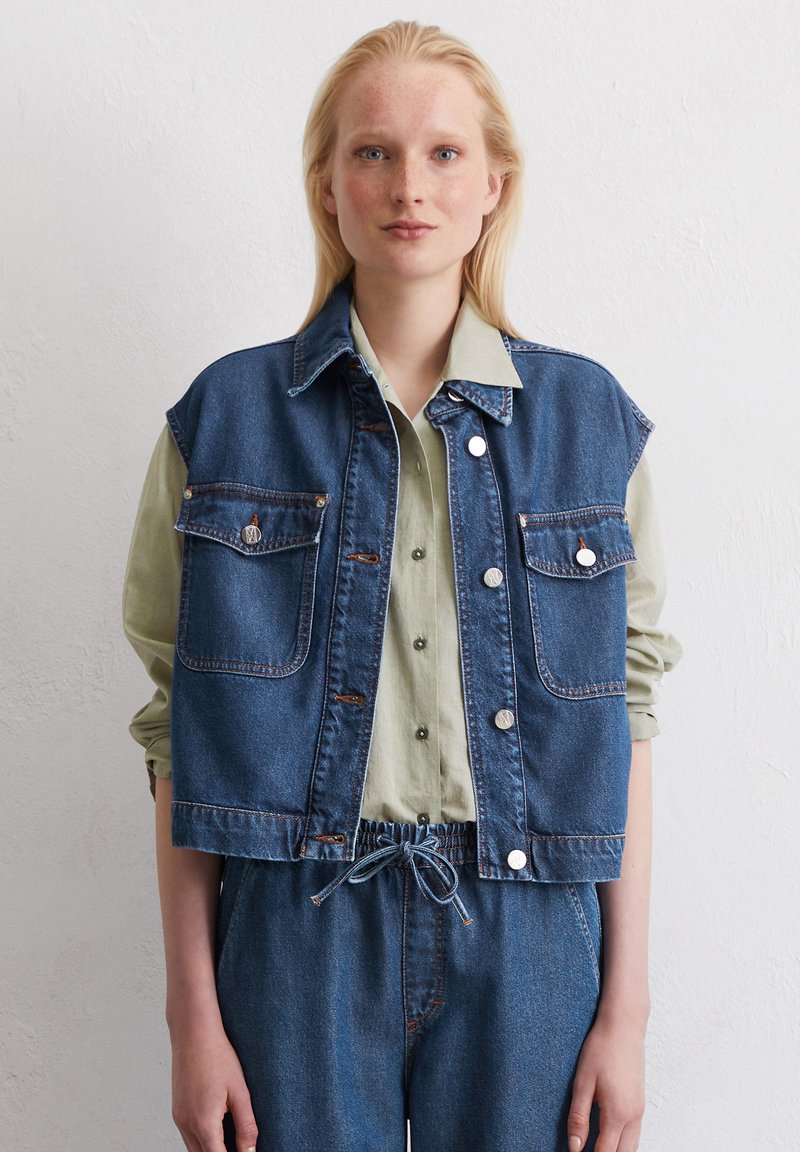 Dunkelblaue Jeansweste mit zwei Fronttaschen, silbernen Druckknöpfen und einem cropped Design, über einem hellgrünen Hemd getragen.