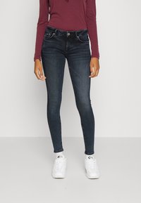 Jeans skinny em denim azul escuro, com uma cintura de média altura, passantes para cinto e cinco bolsos. Combinados com uma blusa de manga longa cor burgundy e sapatilhas brancas.