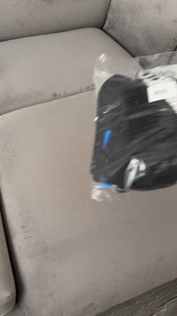 Produto preto embalado em plástico transparente, com um detalhe azul. O fundo apresenta um sofá de textura suave em cinza claro.