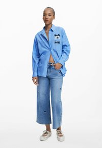 Blauw gestreepte button-up shirt met een zwart grafisch ontwerp, gecombineerd met wijde blauwe denim jeans met bloemenpatronen en lichte schoenen.