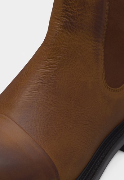 Gros plan sur une botte Chelsea en cuir marron montrant le cuir texturé, le panneau latéral élastique et une partie de la semelle en caoutchouc noire.