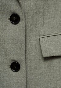 Primo piano di un tessuto grigio testurizzato di una giacca con bottoni neri e un dettaglio di tasca a toppa. La trama è fine, creando un aspetto liscio.