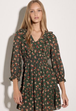 Jeune femme aux longs cheveux blonds porte une robe verte à motif floral avec des volants et des manches longues, debout devant un fond clair uni.