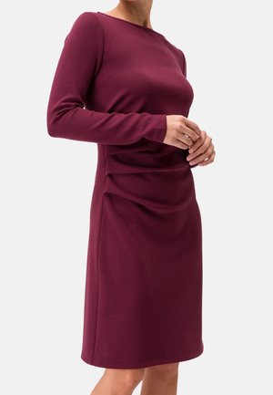 Femme portant une robe bordeaux ajustée, manches longues, longueur genou, avec des détails froncés à la taille, debout devant un fond blanc.