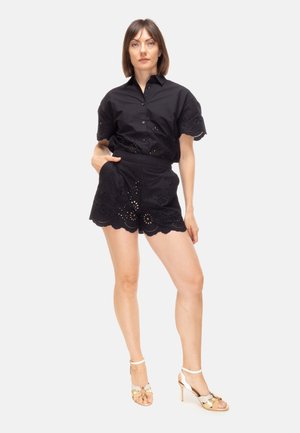 Donna in piedi che indossa una camicetta nera a maniche corte con occhielli e pantaloncini a vita alta abbinati con sandali con tacco alto dorati e bianchi.