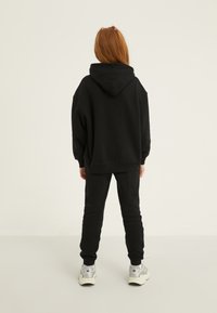 Melna pārāk liela kapucojuma džempera kombinācija ar melniem sporta biksēm. Apģērbu raksturo kokvilnas tekstūra, un tas ir papildināts ar sudraba sporta apaviem.