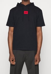 Schwarzes, kurzes T-Shirt mit Kapuze und kurzen Ärmeln, das mit einem roten rechteckigen Aufnäher mit dem "HUGO"-Logo versehen ist. Kordelzugdetails am Halsausschnitt. Baumwollstoff.