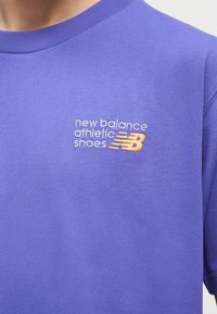 Lilla skjorte med broderet tekst "new balance athletic shoes" og gul "NB" logo på den øvre del af brystet.