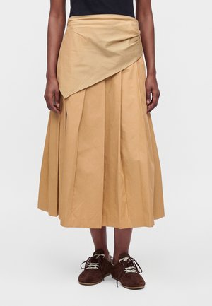 Personne portant une jupe midi plissée beige avec un superposé asymétrique et des chaussures marron foncé à lacets, debout devant un fond uni.