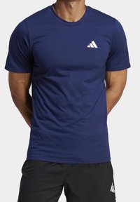 Muž oblečený v navy modrém tričku Adidas s bílým logem na hrudi a černých šortkách Adidas, stojí s rukama za zády.