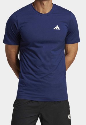 Mężczyzna w granatowej koszulce Adidas z białym logo na piersi oraz czarnych szortach Adidas, stojący z rękami za plecami.
