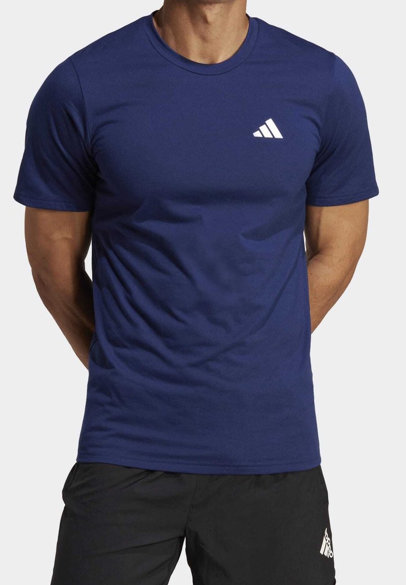 Muž oblečený v navy modrém tričku Adidas s bílým logem na hrudi a černých šortkách Adidas, stojí s rukama za zády.