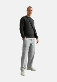 Donkergrijze ribgebreide trui, lichtgrijze cargobroek met zakken en beige sneakers. Het onderwerp draagt een kleine schoudertas.