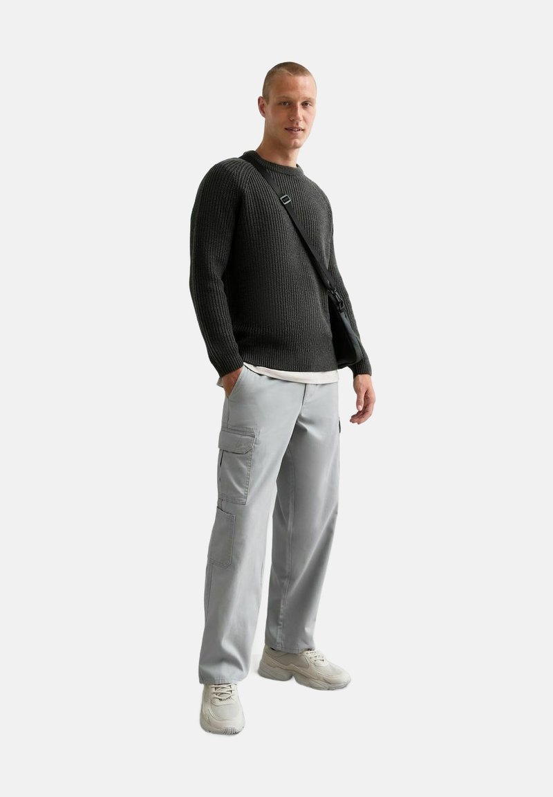 Donkergrijze ribgebreide trui, lichtgrijze cargobroek met zakken en beige sneakers. Het onderwerp draagt een kleine schoudertas.