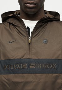 Κοντινό πλάνο ατόμου που φορά καφέ ζακέτα Nike Shox με φερμουάρ στο μισό μήκος, μαύρη ρίγα, μικρό λογότυπο και κεντημένο το σήμα Nike.