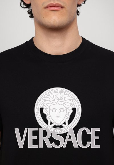 Sort bomulds T-shirt med et stort hvidt Medusa emblem og teksten "VERSACE" i fed hvid skrifttype under grafikken. Cirkulær græsk nøgle kant.