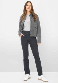 Giacca grigia con texture e bottoni davanti sopra una camicia grigia chiaro, abbinata a pantaloni neri elasticizzati e sneakers bianche. Outfit semplice e informale.