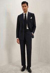 Costum bleumarin cu două rânduri de nasturi, fabricat dintr-un material texturat, cămașă albă, cravată cu buline și pantofi negri din piele; buzunare accentuate în sacou.