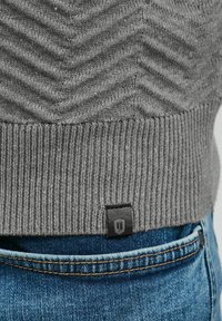 Jersey gris de punto con un patrón de chevrón, dobladillo acanalado y una pequeña etiqueta de logo negra en la parte inferior, combinado con jeans de mezclilla azul.