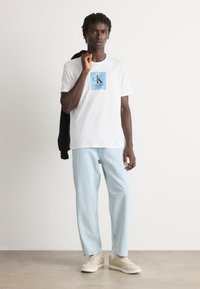 Young man wearing white Calvin Klein T-shirt and light blue jeans, holding a black jacket over his shoulder, standing on a plain floor.