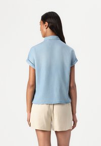 Lyseblå denimskjorte med kort erm, brettet krage, myk tekstur og en lett buet kant, kombinert med beige shortser.