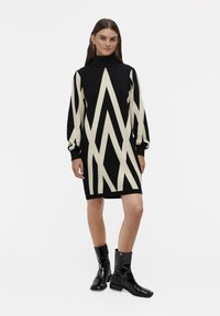 Robe noire tricotée avec un motif géométrique en chevrons crème, col montant et manches longues. Associée à des bottes noires vernies à la cheville.