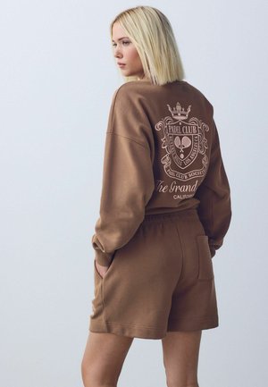 Blonde Frau seitlich gesehen, trägt einen braunen Sweatshirt und passende Shorts, auf der Rückseite des Sweatshirts ist das Emblem "Padel Club" mit Schlägern und Krone.